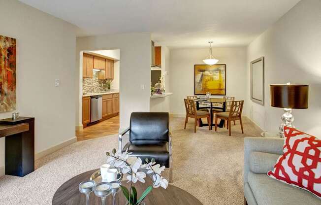 A stylish open living and dining area here at Heritage Hills featuring neutral carpeting, a comfortable sofa with bold accent pillows, and a modern armchair arranged around a round coffee table with decorative accents. The space connects to a dining area with a glass table beneath a pendant light and a kitchen pass-through with warm cabinetry and tile backsplash.