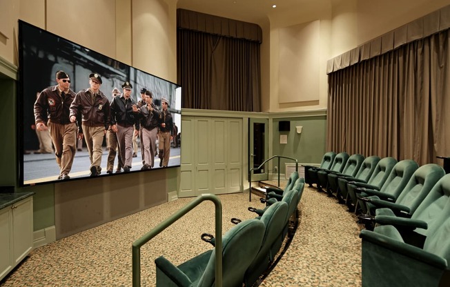 a large screen with a group of men on it in a large room with green chairs