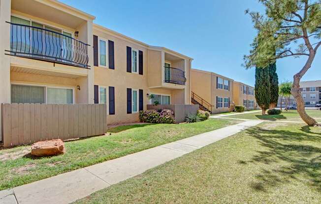 A well-maintained apartment exterior here at Avalon Springs with two-story buildings, private balconies and patios, manicured green lawns, and mature trees lining a paved walkway, creating a calm and welcoming community atmosphere.