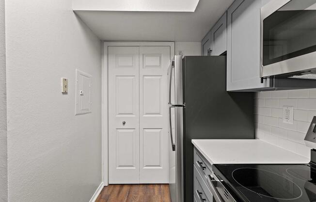 A modern kitchen featuring gray cabinets, a stainless steel refrigerator, and a microwave above a flat-top stove. The space has a white tiled backsplash and a door leading to a closet or pantry. The flooring is wooden, and the walls are painted white, giving the kitchen a bright and contemporary look.