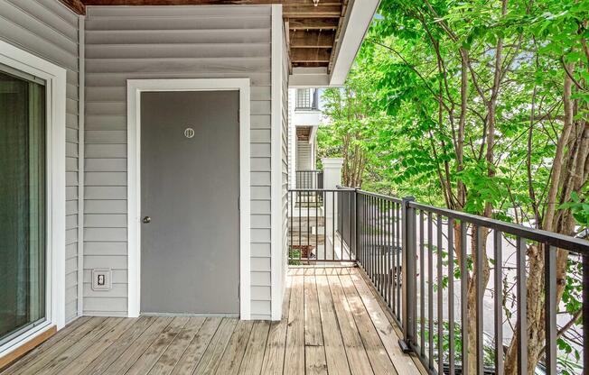 A balcony with wooden flooring, featuring a gray door labeled "11." Lush green trees are visible, adding a natural touch to the space. The house exterior has gray siding, and there is a railing along the balcony's edge. Sunlight filters through the leaves, creating a bright and inviting atmosphere.