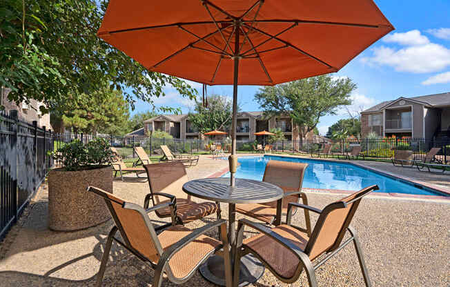 A sunny poolside seating area here at Northridge Court featuring a round outdoor table with four mesh chairs beneath a large orange patio umbrella, set beside a sparkling swimming pool with surrounding lounge chairs, black metal fencing, mature trees, and apartment buildings under a bright blue sky.