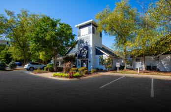 A church with a steeple and a clock tower is surrounded by trees and has a parking lot in front.