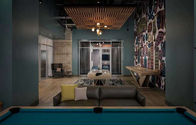 A pool table is in the foreground of a room with a couch, chair, and a patterned wallpaper.