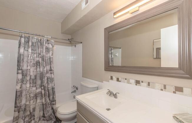 A modern bathroom featuring a shower with a patterned curtain, a white bathtub, a toilet, and a vanity with a sink. The walls are mostly white with a light taupe accent, and there's a large mirror above the sink. Soft lighting is provided by a lamp above the mirror.