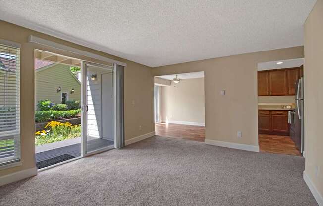 A spacious, empty living room here at Adagio featuring soft beige carpeting, neutral walls, and a wide sliding glass door that opens to a private patio with landscaped greenery outside, while open doorways connect to a bright adjacent room and a kitchen with warm wood cabinets, creating an airy layout with natural light and flexible living space.