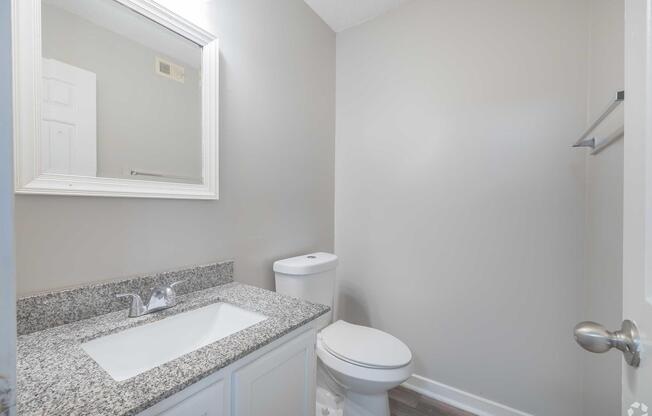 A clean, modern bathroom featuring a granite countertop sink with a mirror above, a white toilet, and a light gray wall. The room is well-lit with natural light coming from an unspecified source, and a towel rack is mounted on the wall. The overall aesthetic is simple and contemporary.