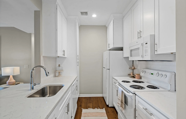 Chic galley kitchen featuring white shaker cabinets, matching white appliances, quartz-style countertops, a stainless steel sink, and wood-look flooring at Kelly’s Ridge Apartments in Columbia, Missouri.