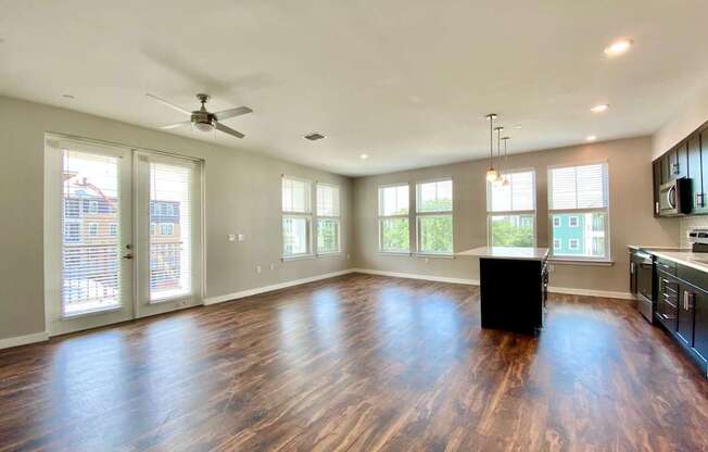 A large open living room with six windows, double doors to a balcony, a ceiling fan, and hardwood-style flooring near a kitchen with an island bar.