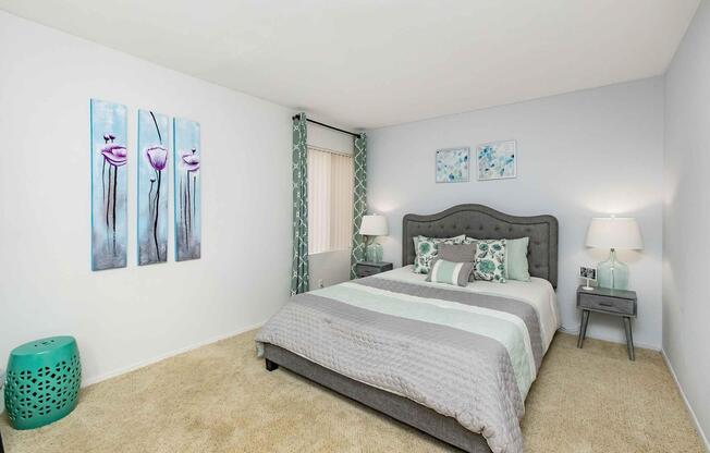 A cozy bedroom featuring a gray upholstered bed with decorative pillows, two bedside tables with lamps, and a vibrant turquoise stool. The walls are adorned with floral artwork, and light curtains frame a window, enhancing the tranquil atmosphere of the space. Soft beige carpeting adds warmth.