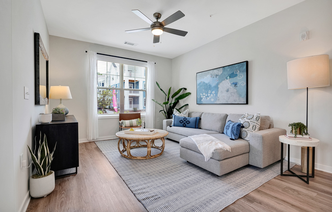 A modern living room featuring a light gray sectional sofa with decorative pillows, a round coffee table, and a tall floor lamp. There is a potted plant in the corner, a console table with a lamp, and large windows letting in natural light. Wall art adds color to the space, creating a cozy atmosphere.