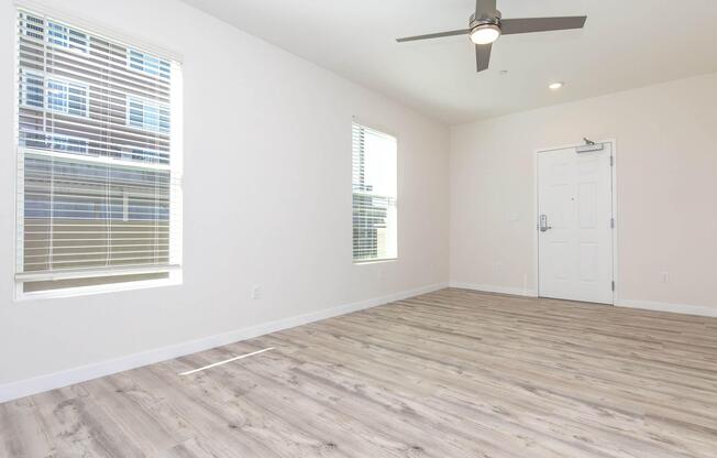 A bright, empty room with light-colored walls and wooden flooring. Two windows allow natural light to enter, with horizontal blinds partially opened. A ceiling fan is mounted on the ceiling, and there is a closed white door on one wall. The space is uncluttered and ready for furnishing.