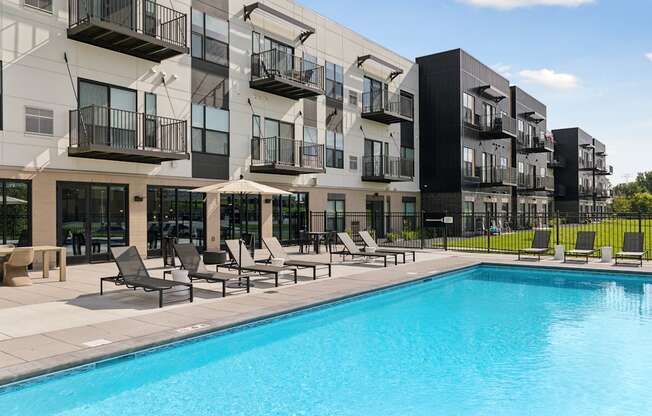 A swimming pool is surrounded by lounge chairs in front of apartment buildings.