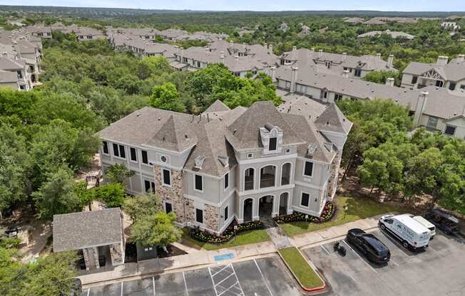 A large house with a grey roof is surrounded by a parking lot and other houses.