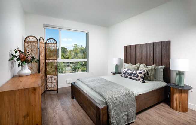 A bedroom with a bed, nightstand, and a window with a view of trees.