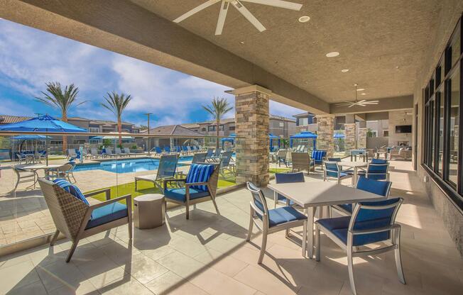 Outdoor seating area with tables and chairs overlooking a swimming pool. Blue umbrellas are set up for shade, and palm trees line the background. The space is well-lit with large windows, creating a relaxed atmosphere for guests.