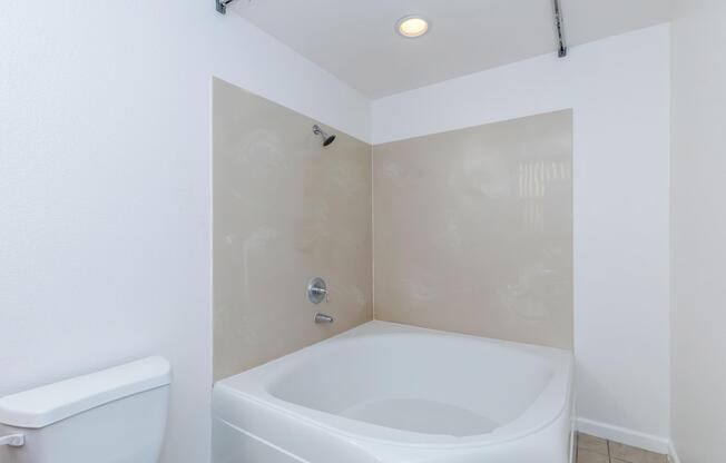 A modern, minimalist bathroom featuring a white bathtub against a beige wall. The bathtub is adjacent to a flush toilet, and there is a shower fixture above the tub. Soft lighting from a ceiling fixture illuminates the space, which has tiled flooring and a clean, uncluttered design.