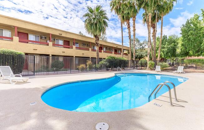 A clear blue swimming pool surrounded by palm trees and neatly landscaped greenery, with lounge chairs positioned nearby. In the background, a two-story building with red accents and multiple windows is visible under a partly cloudy sky.
