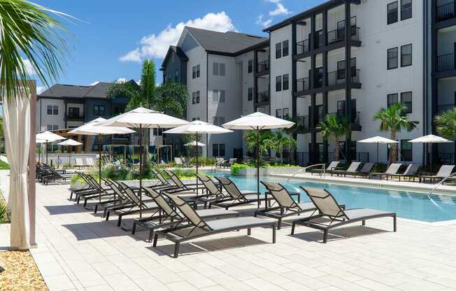A pool area with sun loungers and umbrellas in front of apartment buildings.
