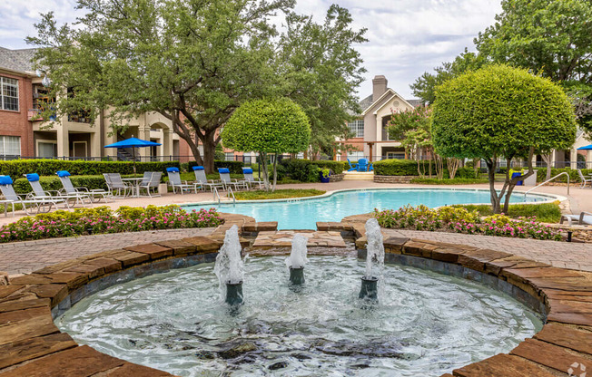 a fountain in front of a swimming pool with chairs and trees