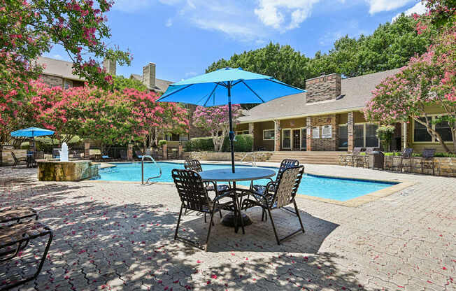 A poolside table with chairs and umbrellas is surrounded by a pool and trees.