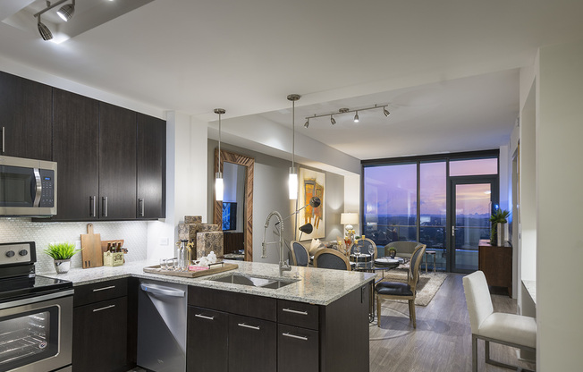 The same open-concept apartment with a pink, purple, and orange city sunset view through the full-width floor-to-ceiling window. The space features ash-gray wood-style floors, dark modern cabinets, stainless steel appliances, granite countertops, and a spacious living area.