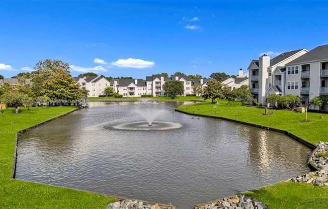 A fountain in the middle of a pond surrounded by grass and buildings.