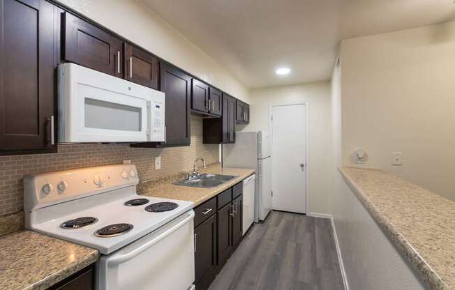 A kitchen with dark wood cabinets and white appliances in a 1 bedroom, 1 bath C, 653 sq ft apartment home with hardwoodlike flooring throughout at The Biltmore Apartments located in the Vickery Midtown neighborhood of Dallas, TX.