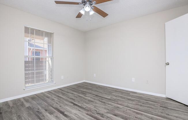 Empty room with light-colored walls, a ceiling fan with wooden blades, and a window with blinds allowing natural light. The floor is covered in laminate wood, and there is a white door on the right side. The space appears clean and minimalistic, suitable for various uses.