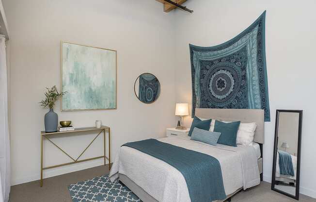 A bedroom with a bed, a side table, a lamp, a mirror, and a painting.