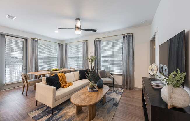 Studio Apartments in Chattanooga, TN - Populus Waterside - Living Room with Luxury Vinyl Tile Flooring, Area Rug, Sofa, Armchair, Coffee Table, Media Console, Wall-Mounted TV, Ceiling Fan, and Large Windows with Blinds.