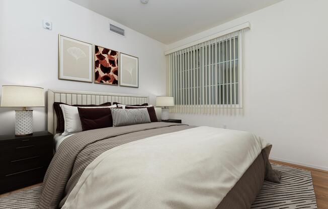 A modern bedroom featuring a neatly made bed with neutral-toned bedding, accented by dark pillows. The wall above the headboard has two framed artworks, and large windows with vertical blinds allow natural light to enter. A textured area rug complements the wooden floor and bedside tables with lamps.