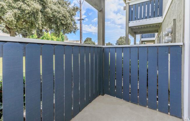 A corner view of a patio enclosed by a blue wooden fence, with a concrete floor. In the background, greenery and a clear blue sky with scattered clouds can be seen, along with a glimpse of a building structure featuring a balcony.