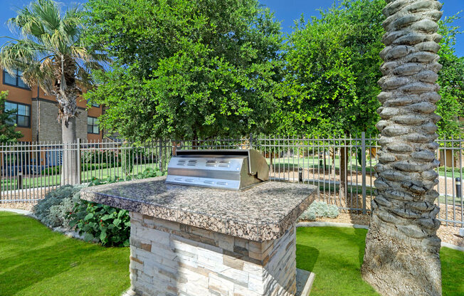 A convenient outdoor grilling area here at Tuscany at Faudree featuring a built-in stainless steel grill set on a stone base, surrounded by green lawn, palm trees, and landscaped greenery. A metal fence and nearby apartment buildings create a relaxed community setting under a clear blue sky.