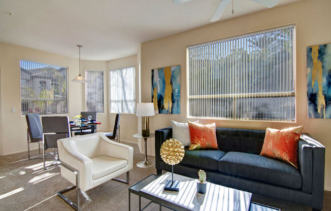 A welcoming living room here at Sonoma Landing featuring a dark upholstered sofa accented with warm orange pillows, a sleek glass coffee table with modern décor, and a white accent chair, while large windows with vertical blinds fill the space with natural light and connect to a dining area with a contemporary table, chairs, and pendant lighting.