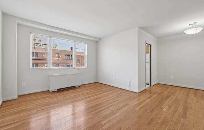 Empty room with wooden floors and a window showing an apartment building outside.