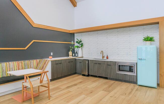A modern kitchen featuring a light blue retro refrigerator, dark wood cabinets, a microwave, and a sink. There's a geometric patterned backsplash and a cozy seating area with colorful cushions. Natural light fills the space, highlighting the warm wood floor and minimalist decor.
