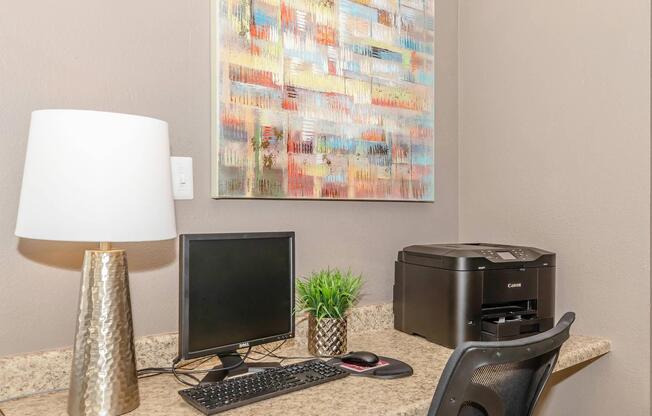 A modern workspace featuring a desktop computer with a monitor, a Canon printer, and a decorative lamp. A small potted plant adds a touch of greenery, and a colorful abstract painting hangs on the wall, providing visual interest to the environment.
