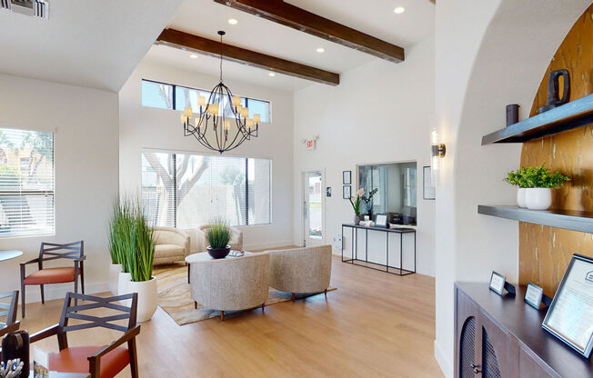 A spacious and airy clubhouse interior here at Pala Mesa featuring soaring ceilings with exposed wood beams, a modern chandelier, and expansive windows that fill the room with natural light. Cozy lounge seating, wood-style flooring, floating shelves with décor, and a welcoming entry area create an inviting and refined community gathering space.