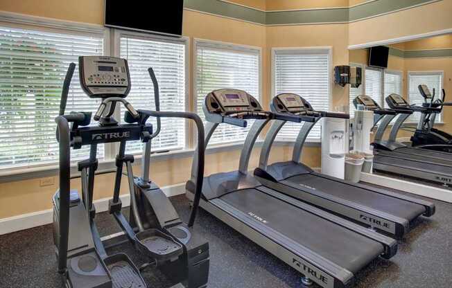 A well-equipped fitness center here at Aspen Creek featuring multiple cardio machines including treadmills and an elliptical trainer, arranged in front of large windows with white blinds that bring in natural light, complemented by mirrored walls, mounted televisions, and rubberized flooring that creates a clean, functional workout space.