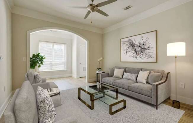A living room with a grey couch, a glass coffee table, and a ceiling fan.