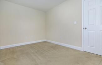 A vacant room with beige walls and light beige carpet. There is a closed white door on the right side, and the room features two electrical outlets on the walls. The space is bright and unfurnished, creating a clean and unadorned atmosphere.
