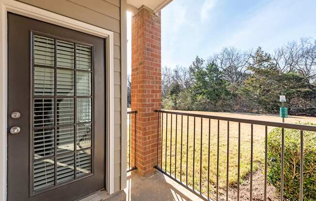 A balcony here at Landing at Mansfield with a dark glass-paneled door, brick column, black metal railing, and a sunny grassy view with trees and shrubs nearby.