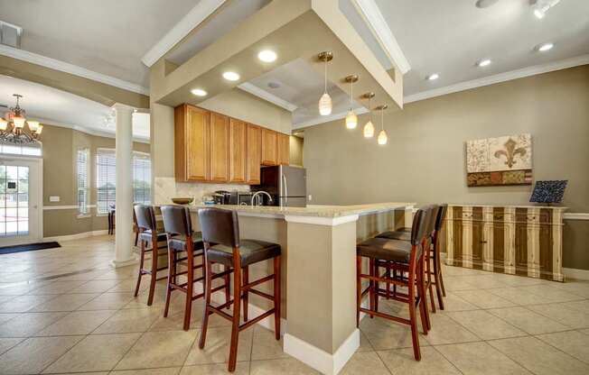 A stylish clubhouse bar and kitchen area here at Mission Green featuring a long granite countertop with barstool seating, warm wood cabinetry, pendant and recessed lighting, tiled flooring, decorative columns, and nearby lounge and dining spaces, creating an inviting setting for resident gatherings, casual dining, and community events.