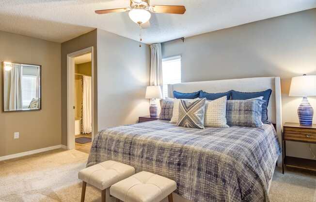 Staged bedroom with a window, gray walls, carpet, bed with seating in front, two nightstands with lamps, ceiling fan, decorative mirror and en-suite bathroom in background.