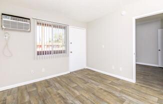 A small, empty room with a light-colored wall, wooden flooring, and a wall-mounted air conditioning unit. There is a window with vertical blinds, an entrance door with a doorknob, and a glimpse of a room through an open door on the right. The space is well-lit and features a minimalist design.
