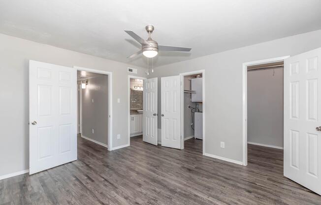 A spacious, modern room featuring light gray walls, a ceiling fan, and hardwood-style flooring. Three open doors lead to different areas, including a bathroom, a closet, and another room. The bright interior creates an inviting atmosphere with ample natural light.