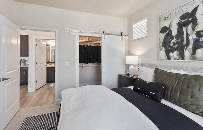 A serene bedroom here at Reflect at Dobson Ranch featuring a plush upholstered bed with crisp white bedding, layered dark accent pillows, and a soft textured rug, complemented by a sleek nightstand with a modern lamp, abstract wall art, and a sliding barn-style closet door, with an open doorway leading to a suite bathroom.