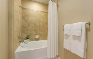 A clean and inviting bathroom here at Mission Green featuring a spacious tub with tiled surround, a crisp white shower curtain, neatly folded white towels on a wall-mounted rack, warm neutral wall tones, and simple decorative accents that create a bright, comfortable, and well-maintained bathing space.