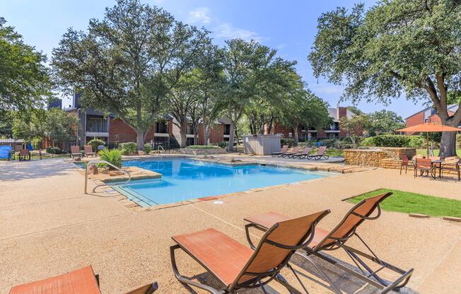 A spacious outdoor pool area surrounded by lounge chairs and trees. The pool is clear and inviting, with a stone border. In the background, there are multi-story residential buildings and additional seating areas, creating a relaxing atmosphere. The sky is blue and sunny.